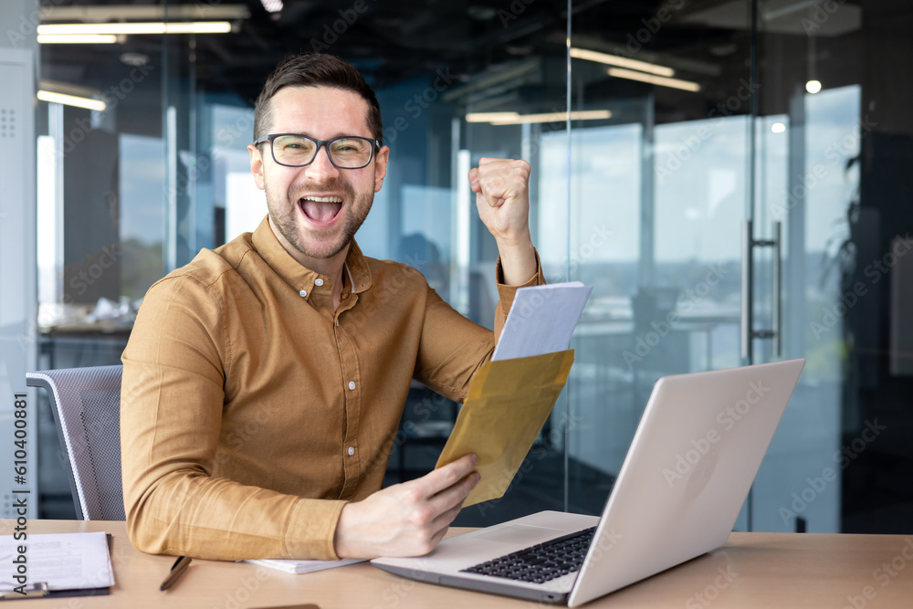 Portrait of a happy young businessman sitting in the office holding an envelope with the documents. A successful financial deal. He looks at the camera, shows a victory gesture with his hand. Portrait of a happy young businessman sitting in the office holding an envelope with the documents. A successful financial deal. He looks at the camera, shows a victory gesture with his hand.