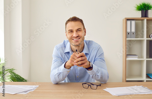 Portrait of happy businessman or remote worker at online business meeting via video conference call. Young man in shirt sitting at office table, holding pen, looking at camera and smiling