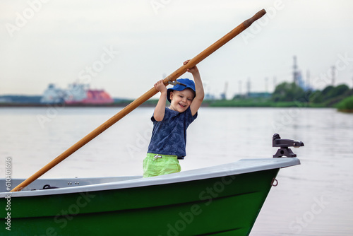 A small cheerful boy is playing in a fishing boat, he is trying to lift a heavy and big oar