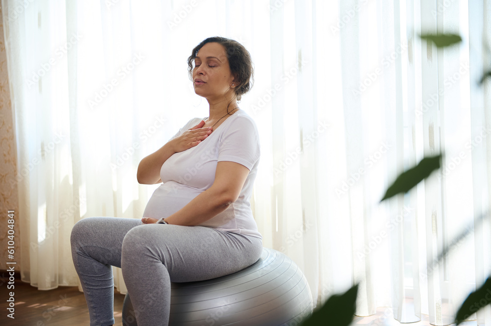 Pregnant woman holding hands on her chest and belly, doing pranayama breathing exercises, sitting relaxed on fitball at home. Spiritual and emotional concept of harmony in pregnancy and maternity time