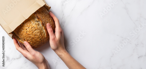 Fresh sourdough bread in woman's hands, top view. Woman takes a loaf of rustic wheat bread from a paper bag. Copy space banner on white marble background.