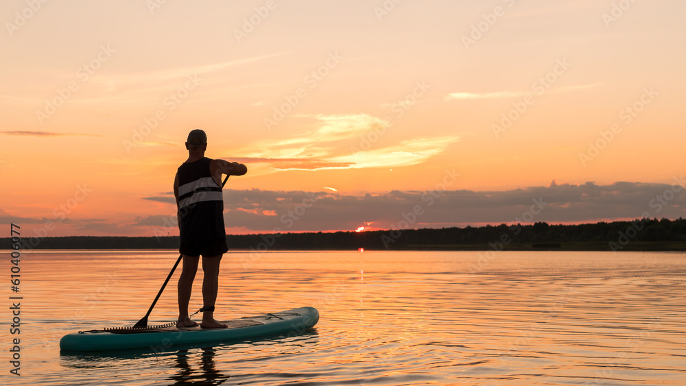 A man in shorts on a SUP board with a paddle at sunset swims in the water of the lake against the backdrop of the sunset sky.