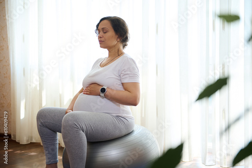 Spiritual and emotional concept of harmony in pregnancy and maternity time. Relaxed conscious pregnant woman holding hands on her belly, doing breathing exercises, sitting relaxed on fit ball at home