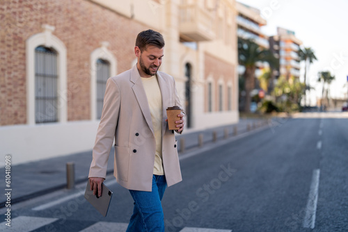 Satisfied businessman holding coffee to go and digital tablet, crossing the street in the city. Photo of male business professional wearing smart casual clothes crossing the street with copy space.