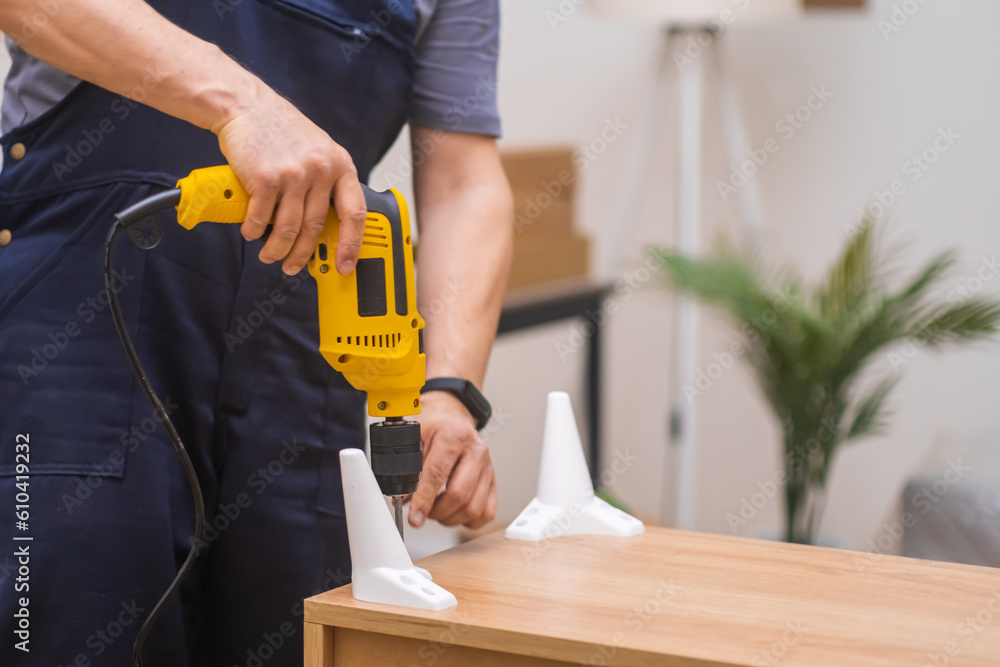 Skilled furniture assembler in uniform completing the assembly process of a wooden rack, attaching legs using an electric screwdriver in a stylish apartment. Skilled furniture assembler in uniform completing the assembly process of a wooden rack, attaching legs using an electric screwdriver in a stylish apartment.