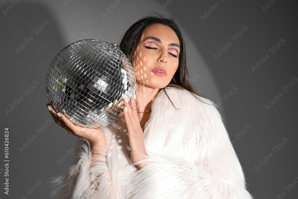 Naklejka premium Fashionable beautiful young woman in fur coat with disco ball on grey background