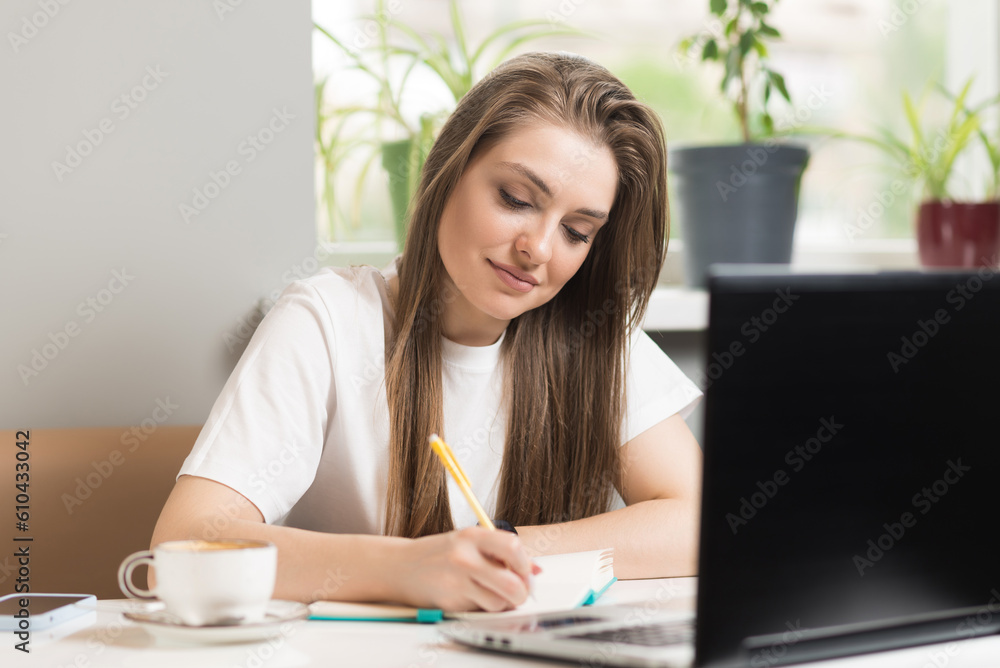 A freelance girl writes in a notebook and uses a laptop in a cafe. Freelance concept. A freelance girl writes in a notebook and uses a laptop in a cafe. Freelance concept.