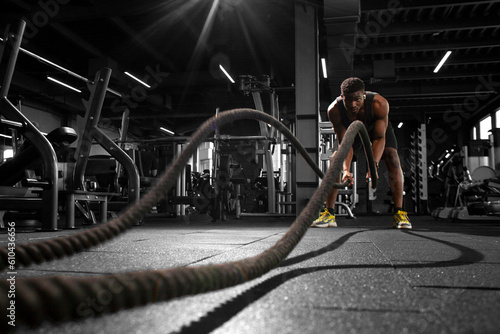 athletic african american man trains with ropes in dark gym, motivated guy waving ropes in fitness club