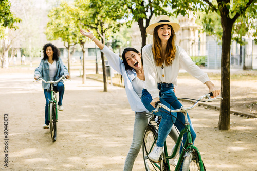 Young group of three multiracial women enjoying bicycle ride in city park. Diverse female friends having fun together riding bike during summer vacation outside. Youth community and travel concept.