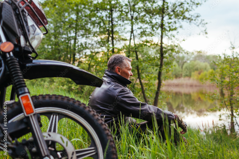 Portrait of senior biker relaxing by motobike outdoors. Man wearing leather jacket enjoying river landscape.
