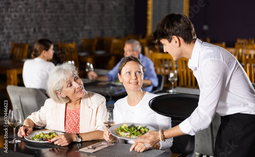 Young guy waiter in uniform serves ordered dish to elderly woman and adult woman in restaurant