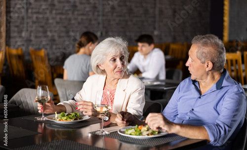 Cheerful elderly couple spending time together in cozy restaurant. Man and woman having fun while talking and enjoying light dinner with wine at table