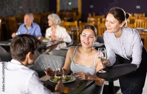 Polite smiling waitress serving ordered drinks to young couple sitting at table  visiting restaurant for romantic dinner