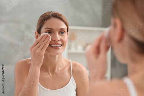 Beautiful woman removing makeup with cotton pad near mirror indoors