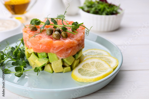 Tasty salmon tartare with avocado, lemon, capers and microgreens on white wooden table, closeup