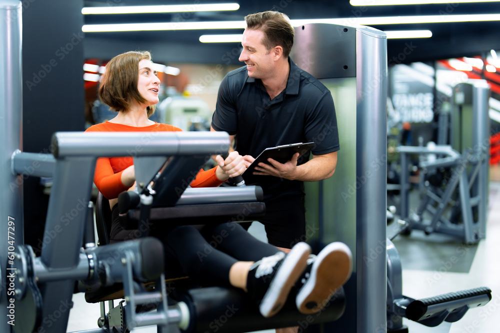 Healthy young woman exercising in fitness gym while supervised by male trainer.