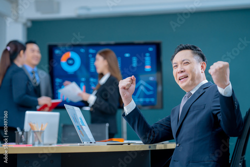 Smiling middle-aged executive asian businessman sitting at a worktable a modern office, typing on computer keyboard, and talk to work, sending emails to his business partners, working on marketing.