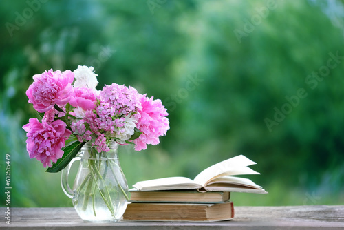flowers bouquet in glass jug and books on table in garden, green natural abstract background. romantic atmosphere nature image. reading time. spring, summer season. template for design. copy space