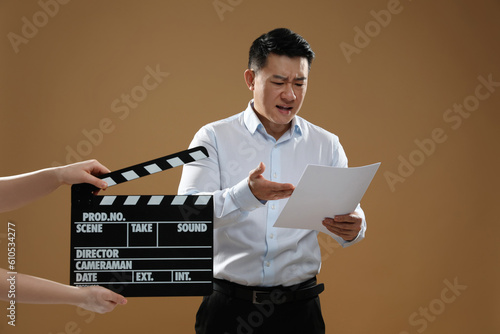Emotional asian actor performing role while second assistant camera holding clapperboard on brown background. Film industry