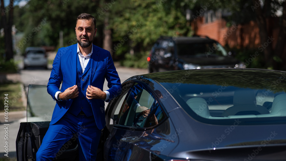 Caucasian bearded man in a blue suit gets out of a black electro car in the countryside in summer.