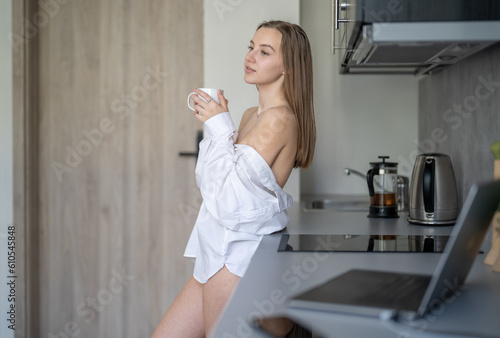 Beautiful Young Girl is Standing in the Kitchen and Drinking Coffee. Morning Routine. Wearing White shirts. Blurry Laptop and Mobile Phone on the Desk.