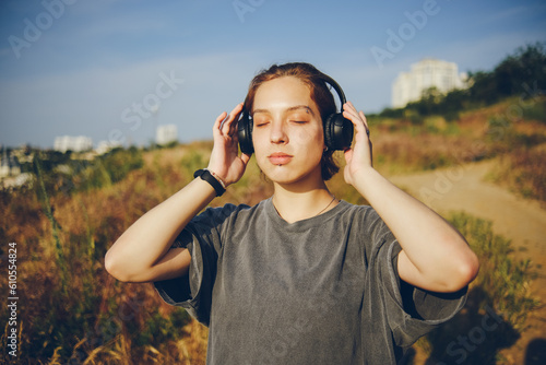 A young girl dreamily listens to music in nature with her eyes closed. The girl is wearing full-size black headphones. Place for text on the t-shirt.