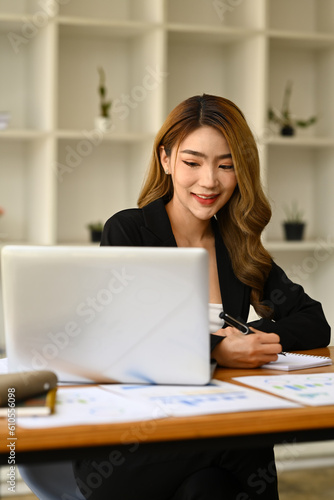 Portrait of beautiful female manager in trendy suit using laptop and making notes on notebook