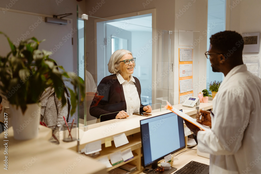 Smiling senior female patient communicating with male receptionist through transparent shield in clinic Smiling senior female patient communicating with male receptionist through transparent shield in clinic
