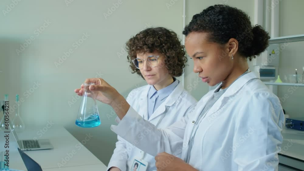 Closeup of two diverse lab scientists looking at flask with blue liquid having discussion in laboratory at daytime Closeup of two diverse lab scientists looking at flask with blue liquid having discussion in laboratory at daytime
