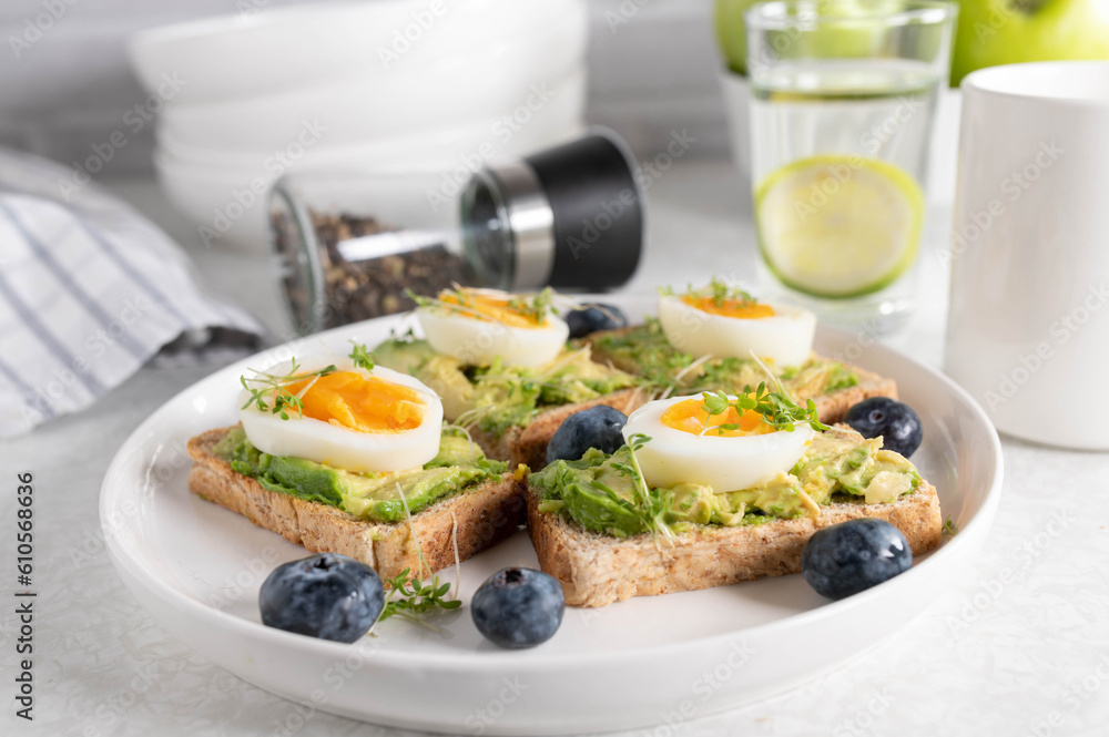 Breakfast toast with avocado spread, boiled eggs and cress on a plate. Served with a glass of water and coffee on a kitchen table.