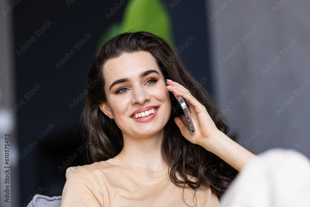 Fototapeta premium Happy woman talking on mobile phone sitting on comfortable couch at home