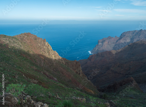 View of illuminated sharp rock and cliffs on top of La Merica mountain with cacti, pine and palm trees. Camino La Merica hiking trail. Arure, Valle Gran Rey , La Gomera, Canary Islands, Spain. photo