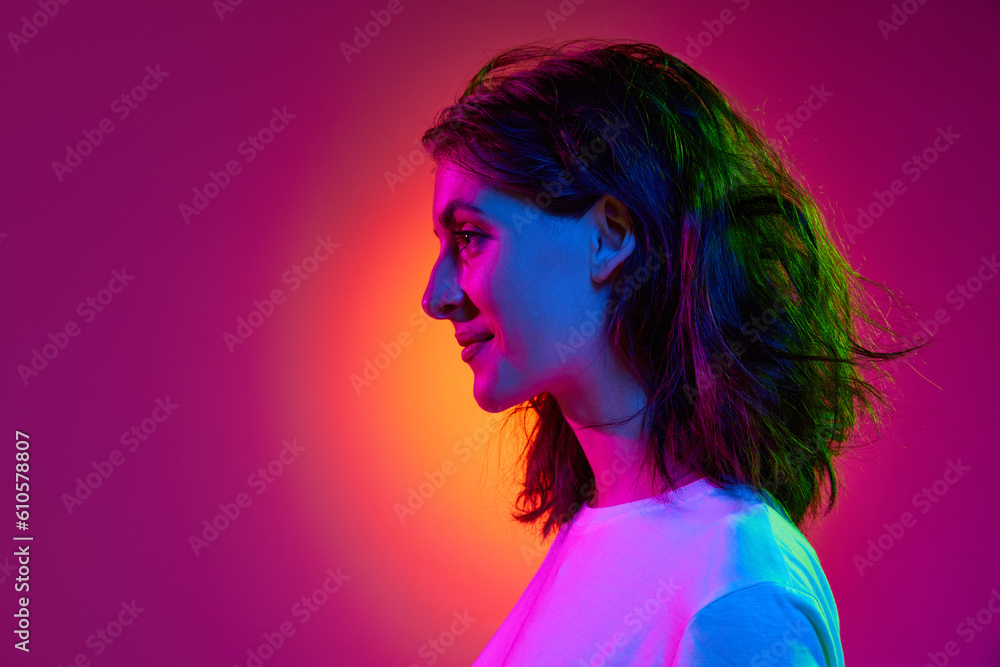 Portrait of young charming girl, student with wavy hair posing over pink studio background in neon light. Side view. Cheerful mood