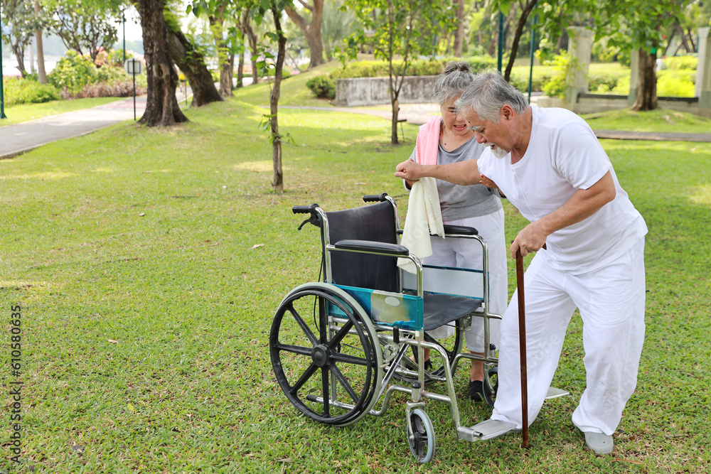 Asian senior woman or caregiver helping and consoling senior man walk with wheelchair at park outdoor. Elderly wife taking good help care and support of elder husband patient outside the house. Asian senior woman or caregiver helping and consoling senior man walk with wheelchair at park outdoor. Elderly wife taking good help care and support of elder husband patient outside the house.