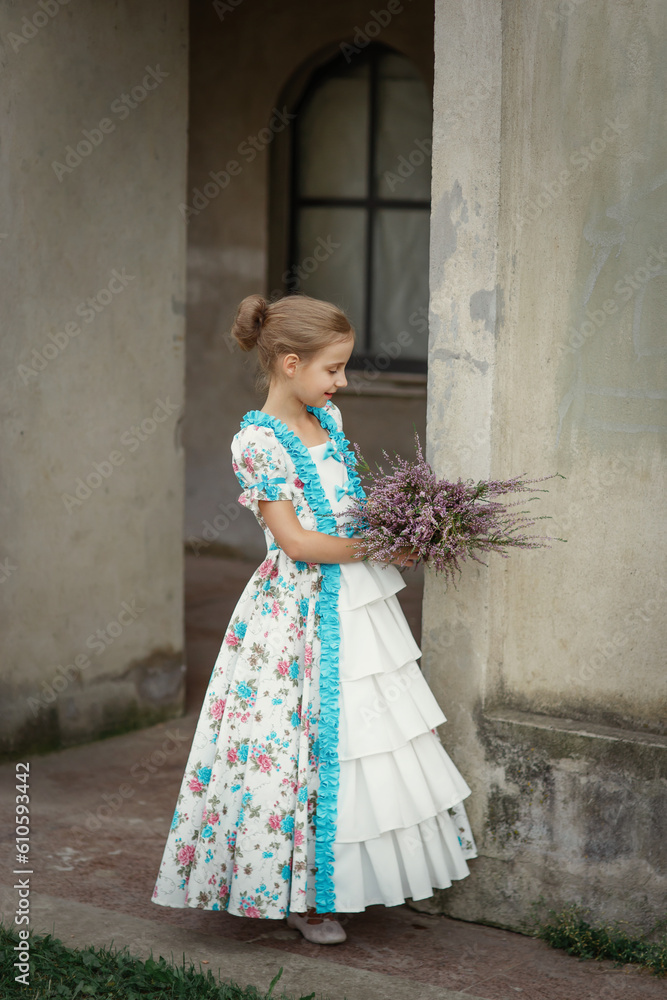 Young cute girl in vintage elegant old-fashioned dress standing sideways with bouquet of heather flowers in front of old mansion in countryside, summer retro kid's portrait in village provence style