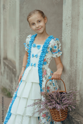 Young cute smiling girl in vintage elegant dress standing with basket of heather flowers near wall of old mansion in countryside looking at camera, summer outdoor story  in provence retro style