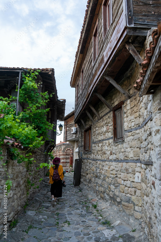 Woman photographer standing on an alley with rock house and traditional footpath in Bulgaria with typical architecture of the building with wood. Village Downtown female tourist walking with a hat and