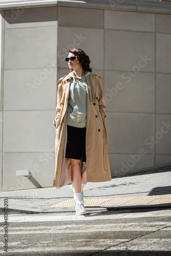 Wallpaper Mural full length of trendy woman with wavy brunette hair crossing road in beige trench coat, grey hoodie, black shorts, white sneakers and dark sunglasses near modern building in city Torontodigital.ca