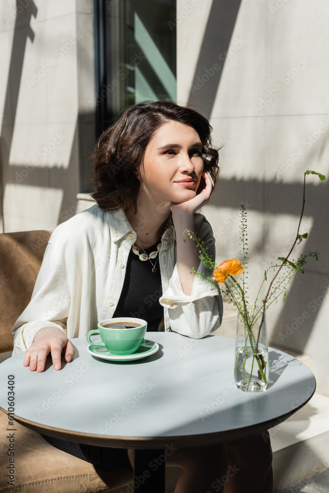 smiling woman sitting at white round table near cup of black aromatic coffee, saucer, glass vase with yellow rose and green plants on summer terrace of hotel cafe in morning sunlight smiling woman sitting at white round table near cup of black aromatic coffee, saucer, glass vase with yellow rose and green plants on summer terrace of hotel cafe in morning sunlight