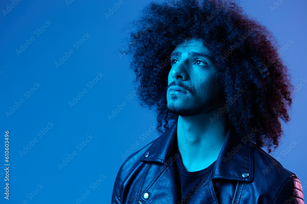 Portrait of fashion man with curly hair on blue background multinational, colored light, black leather jacket trend, modern concept.