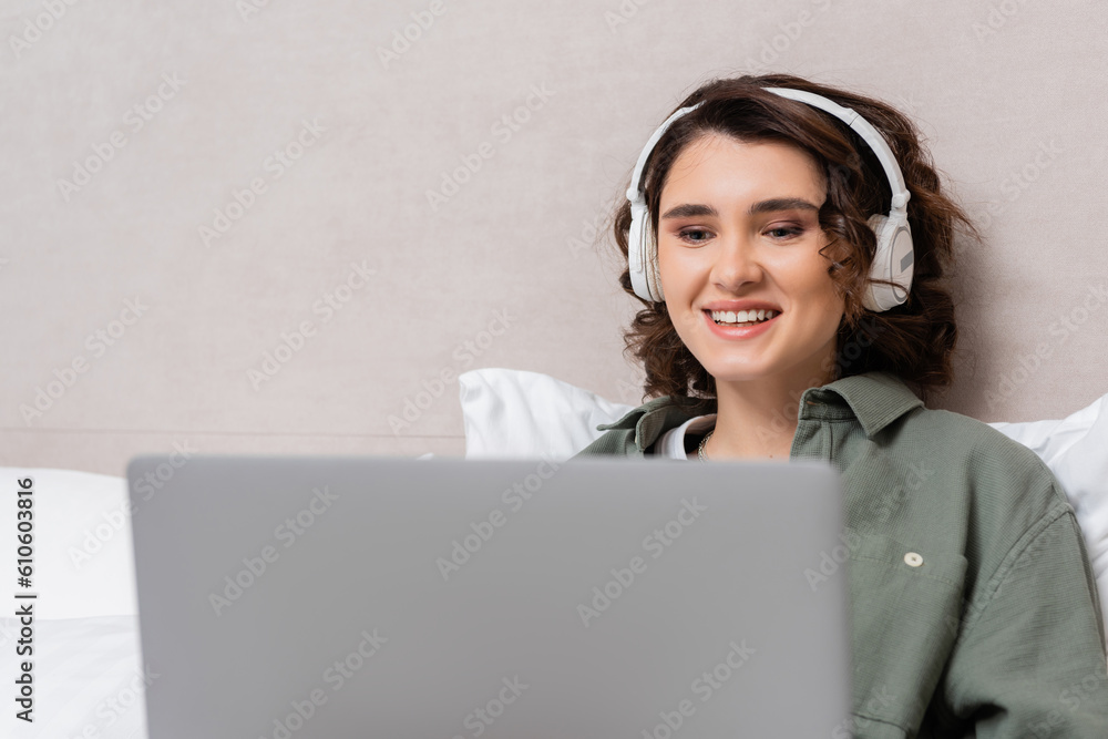 young and cheerful woman with wavy brunette hair watching film on blurred laptop while sitting in wireless headphones near grey wall and white pillows in modern hotel room young and cheerful woman with wavy brunette hair watching film on blurred laptop while sitting in wireless headphones near grey wall and white pillows in modern hotel room