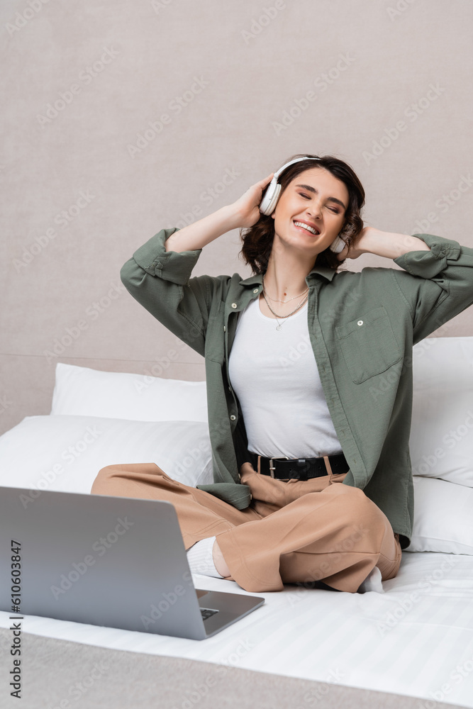 carefree woman with wavy brunette hair and closed eyes listening musical podcast in wireless headphones while sitting on bed near laptop and white pillows in hotel suite, leisure and travel carefree woman with wavy brunette hair and closed eyes listening musical podcast in wireless headphones while sitting on bed near laptop and white pillows in hotel suite, leisure and travel