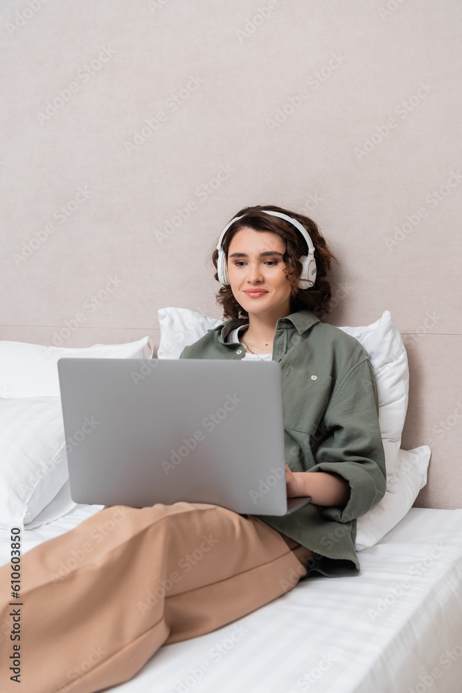 young woman with wavy brunette hair, in casual clothes and wireless headphones sitting on bed near white pillows and smiling while watching film on laptop in hotel room young woman with wavy brunette hair, in casual clothes and wireless headphones sitting on bed near white pillows and smiling while watching film on laptop in hotel room