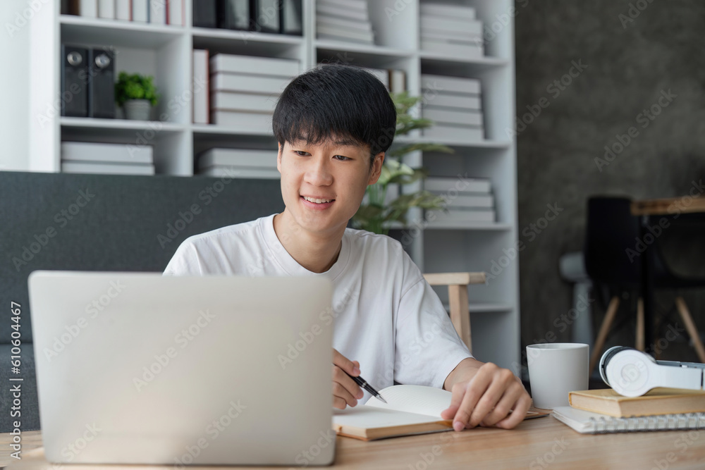 young man studying at home using his laptop, receiving lectures online and taking notes, smiling young man studying at home using his laptop, receiving lectures online and taking notes, smiling