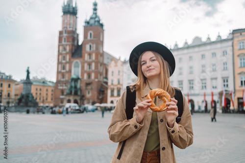 Happy young blonde female tourist with stylish clothes and a boho hat eating bagel obwarzanek traditional polish cuisine snack waling on Market square in Krakow. Traveling Europe. St. Marys Basilica photo