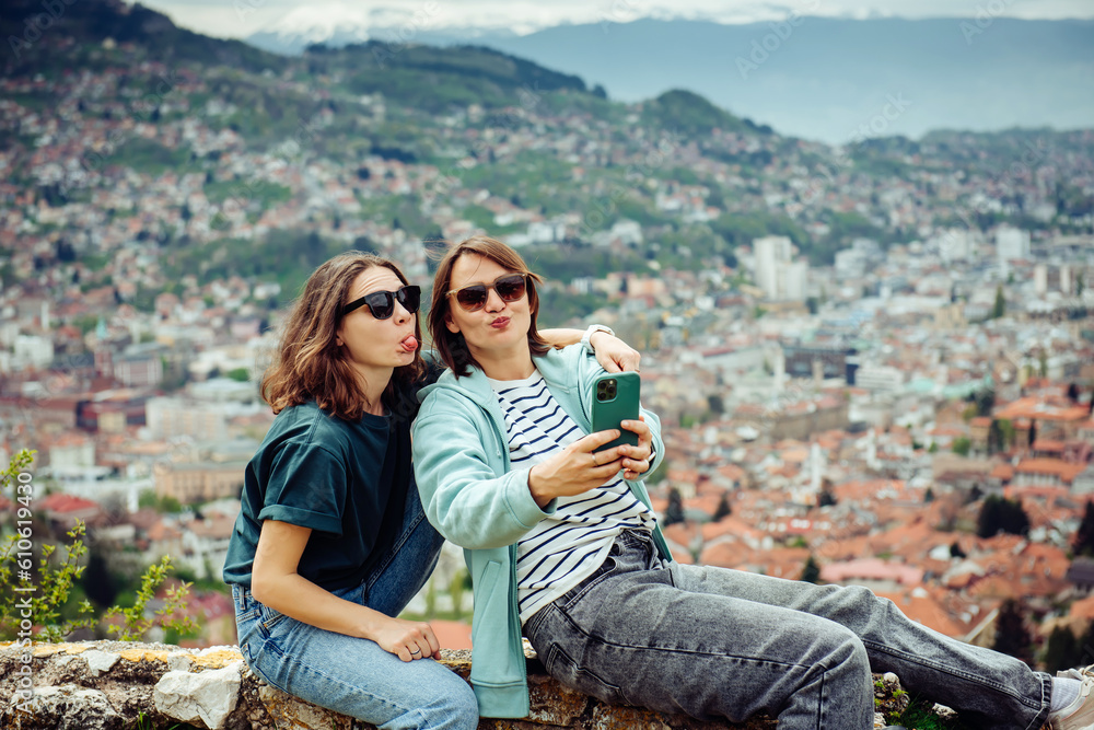 Two cheerful traveler girls in dark glasses are having fun and taking pictures against the background of Sarajevo, a beautiful European city in the mountains Two cheerful traveler girls in dark glasses are having fun and taking pictures against the background of Sarajevo, a beautiful European city in the mountains