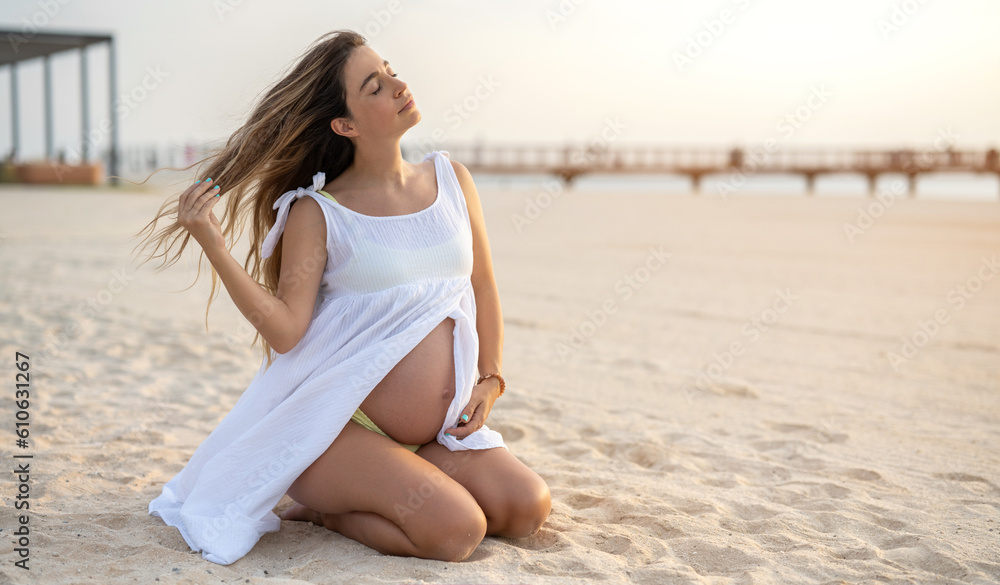 Beautiful young pregnant woman sitting on her knees enjoying beautiful moments on the beach during sunset.