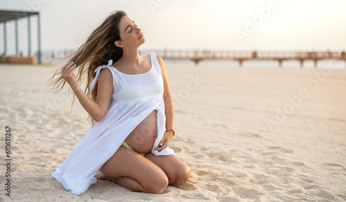 Fototapeta Naklejka Na Ścianę i Meble -  Beautiful young pregnant woman sitting on her knees enjoying beautiful moments on the beach during sunset.