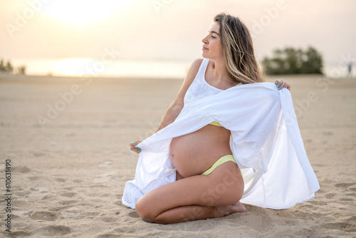 Young woman wearing white dress sitting on her kneed showing belly and looking over sunset on the beach.
