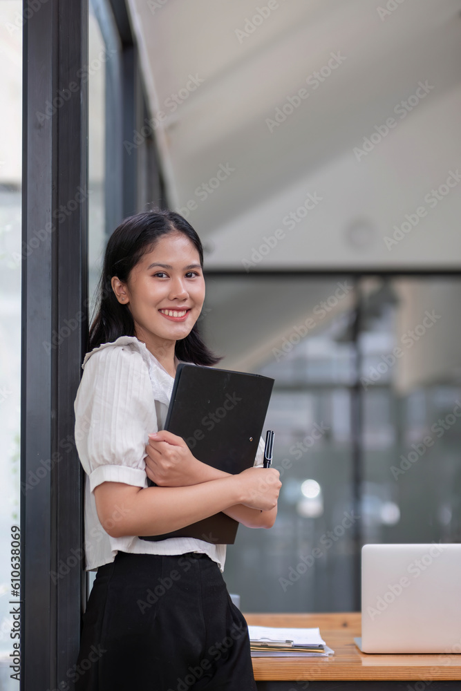 Charming young Asian businesswoman with a smile standing holding document clipboard at the office. Looking at camera. Charming young Asian businesswoman with a smile standing holding document clipboard at the office. Looking at camera.